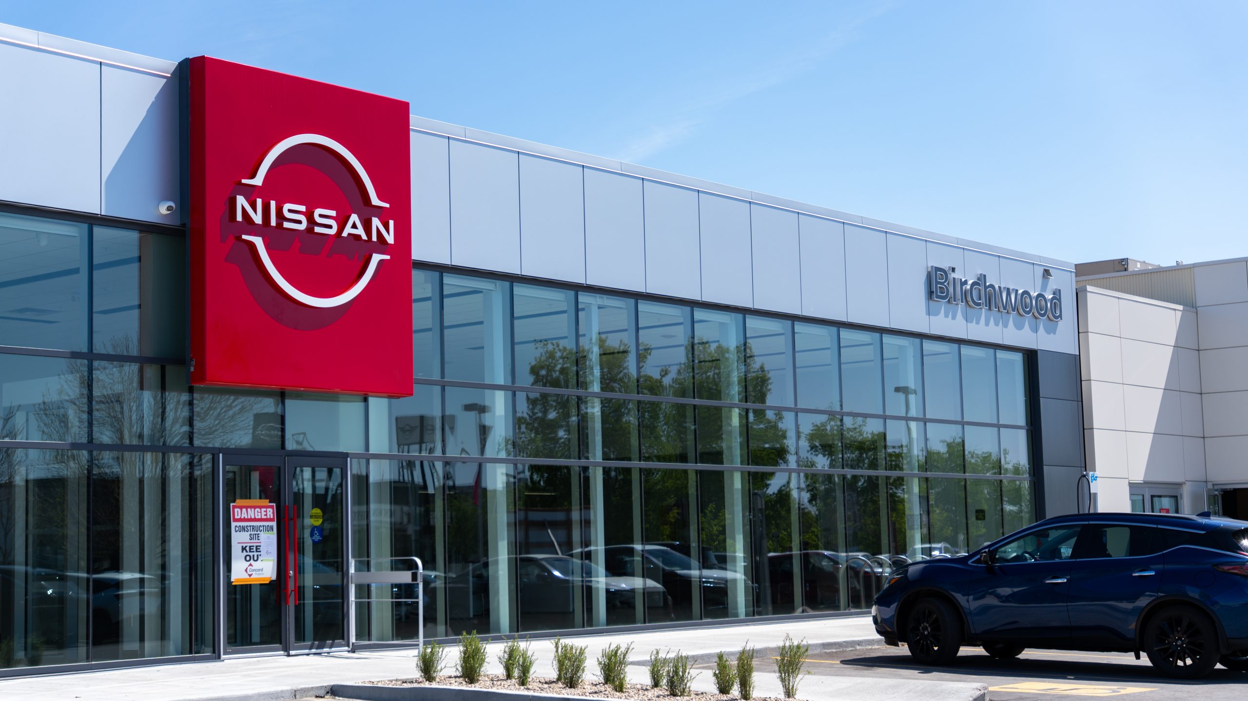 Exterior of Birchwood Nissan dealership with large red Nissan sign, modern glass façade, and a dark blue Nissan SUV parked in front.